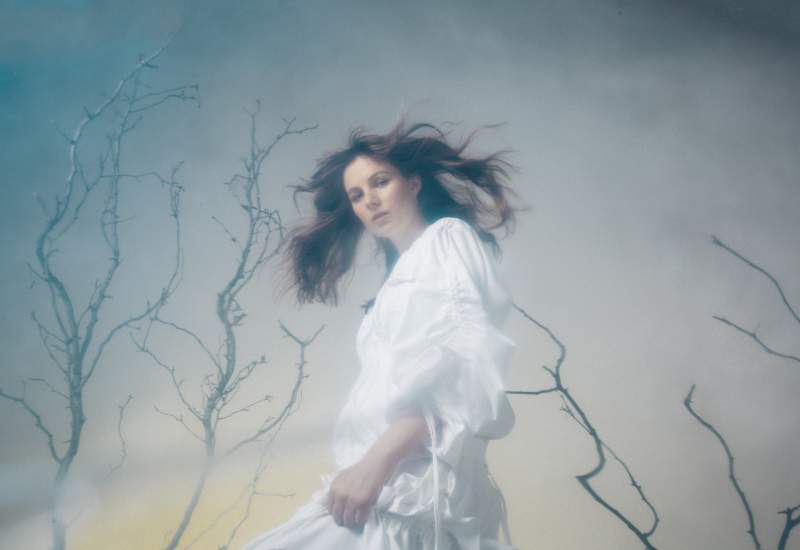 Gena Rose Bruce standing outside with windswept hair. They are wearing a flowing white gown.