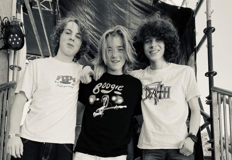 Three members of Cheeky Geezers smiling and standing on steps leading up to a stage