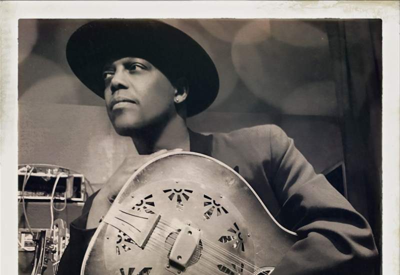 A sepia photo of Eric Bibb holding a guitar and looking off to the right.