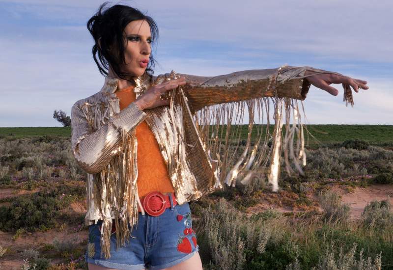 Adalita wearing a gold tasseled jacket with their arms up and hair tied back.