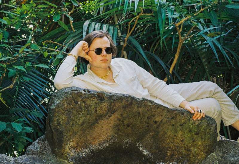 Milo laying down on a large grey rock in front of a background of deep green plants. He is outdoors 