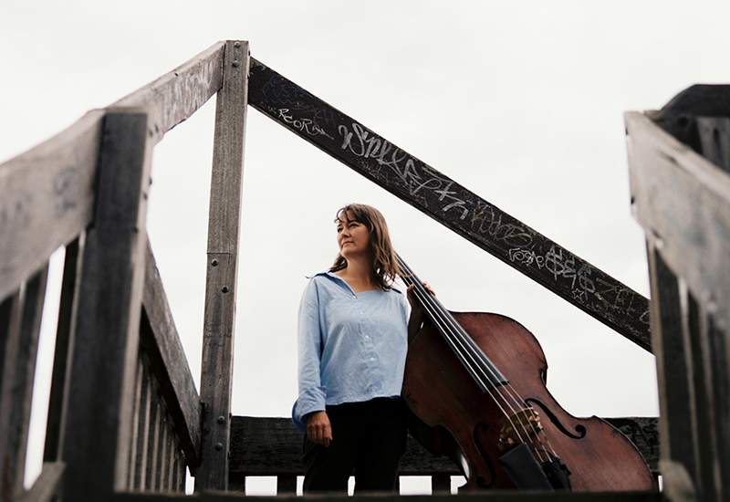 Tamara standing outside at the top of a staircase with their double bass.